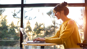 jonge vrouw in gele trui met laptop voor het raam